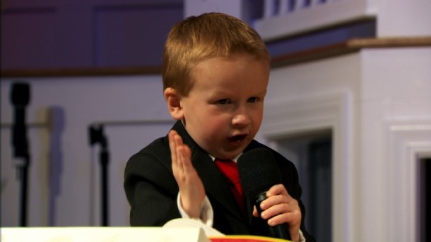 Criança de terno se comunicando com um microfone na mão